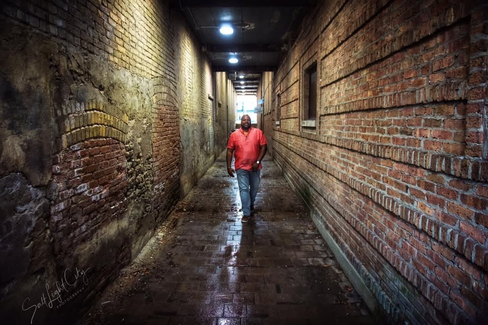 Person in red jacket walking alone through narrow wet brick alley at night with overhead lights